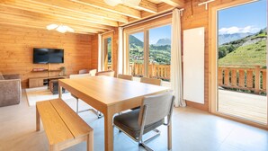 A wooden interior with a large dining table, chairs, a bench, a TV, and large windows offering a mountain view.