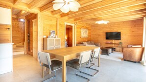 A wooden interior with a dining table, chairs, a sofa, a TV, and a staircase.