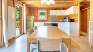 A wooden cabin with a dining table, chairs, a kitchen with white cabinets, and a window overlooking greenery.