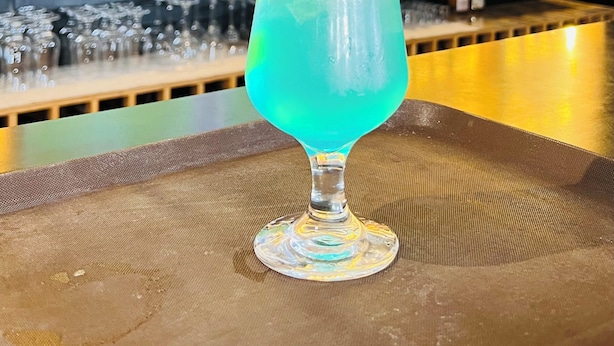Baaritiskialue, jossa on suuri näyttö, jolta näkyy urheilutapahtuma, hyllyt täynnä alkoholijuomapulloja sekä lasi sinistä cocktailia, joka on koristeltu limenviipaleella.