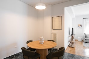 A modern living room with a round wooden dining table, four dark chairs, a white vase, a framed menu on the wall, a radiator, a TV, and a grey sofa. - LIV - Premium Pagrati Apartment With View (Athens)