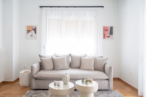 A modern living room with a beige sofa, two white side tables, and a large window with sheer curtains. - LIV - Premium Pagrati Apartment With View (Athens)