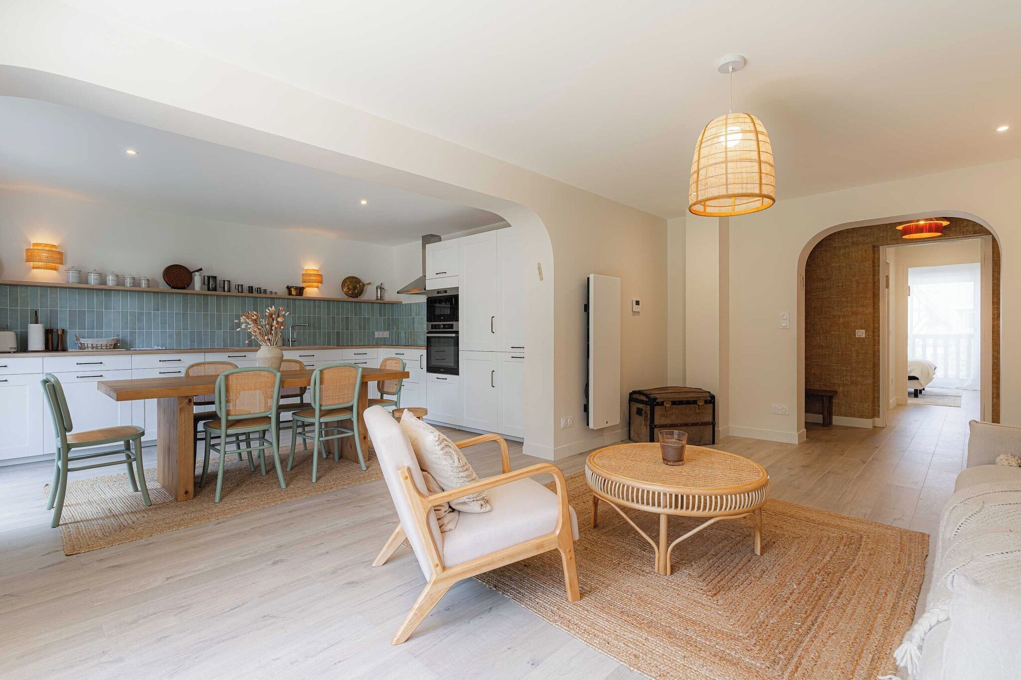 Eine moderne Küche mit weißen Schränken, eine Essecke mit rundem Tisch und Stühlen sowie ein Wohnzimmer mit einer Couch und einer Lampe.