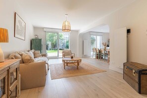 Ein geräumiges Wohnzimmer mit einer Couch, einem runden Tisch und einem Korbstuhl. Ein großes Fenster bietet Blick ins Freie.