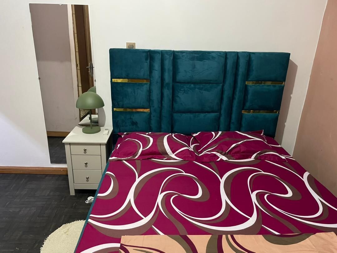 Un dormitorio con cabecera color turquesa, colcha estampada, lámpara de noche y mesita de noche pequeña.