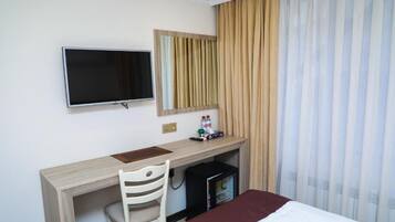 A hotel room with a bed, a desk with a chair, a television, a mirror, a minibar, and curtains.