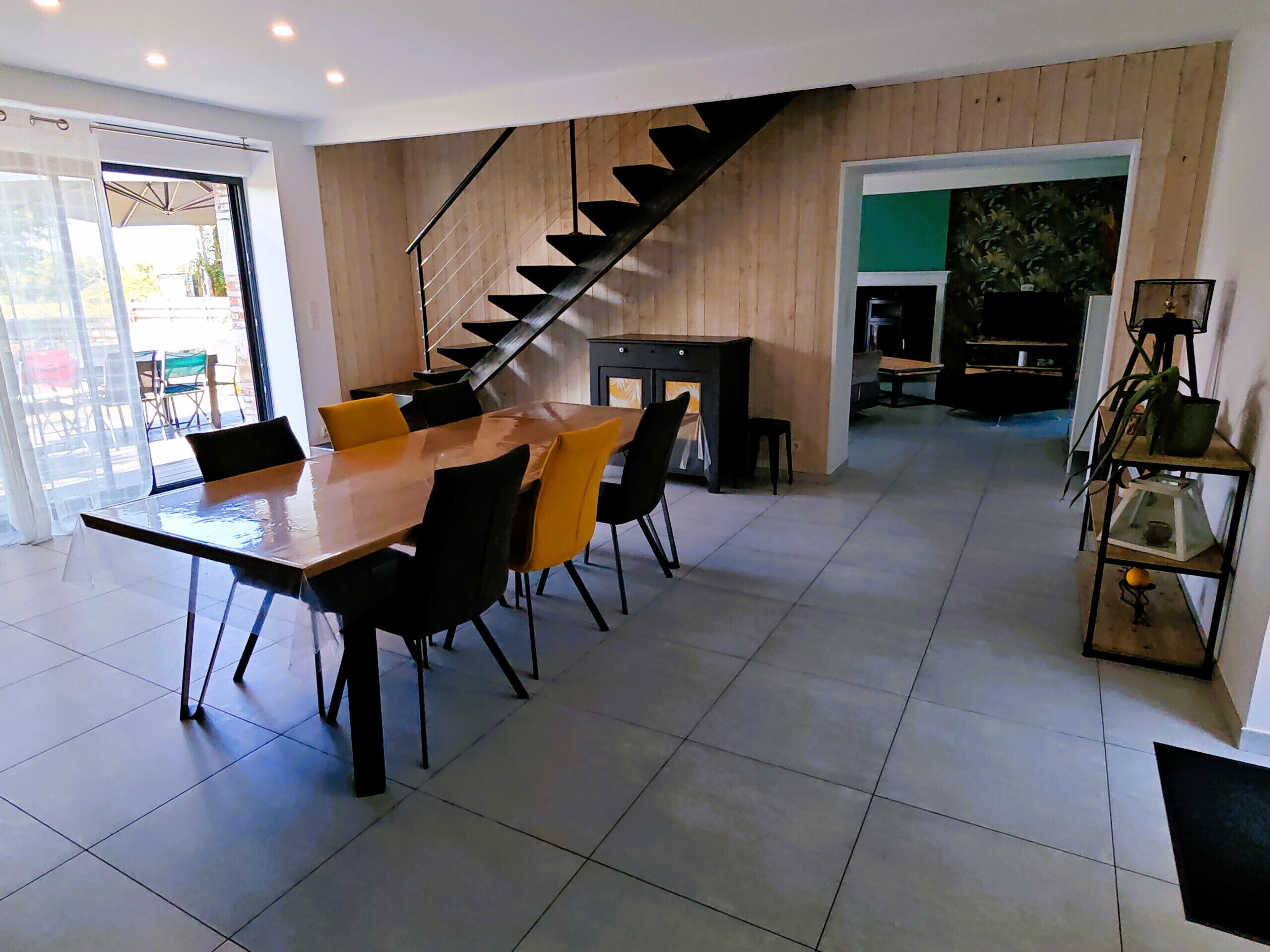 A modern kitchen with a wooden staircase, a dining area with a large table and chairs, and a view of the outdoors through a glass door.