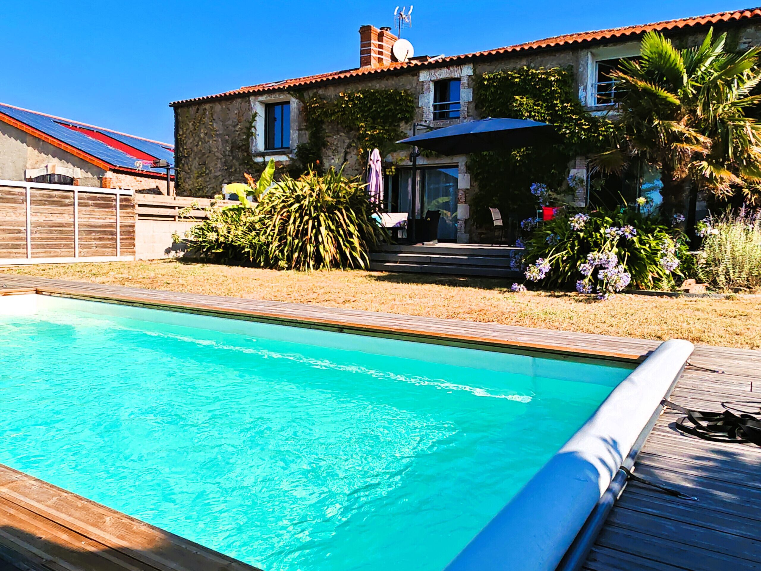 A swimming pool with a wooden deck, a stone house with a red roof, and a garden with various plants and flowers.