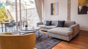 Una sala de estar moderna con un sofá gris, una mesa de centro redonda de vidrio y un sillón amarillo. Hay un ventanal con vistas a un edificio.