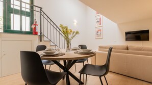 A modern dining area with a round table, black chairs, and a vase of flowers. A sofa and a television are visible in the background.