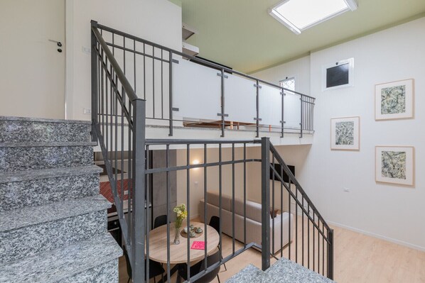 A staircase with metal railings leading to a loft area, a round table with a vase and a pink book, and framed artwork on the walls.