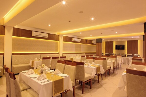 Une salle à manger d’hôtel avec de nombreuses tables dressées pour les clients, des climatiseurs fixés aux murs et un téléviseur accroché au mur.