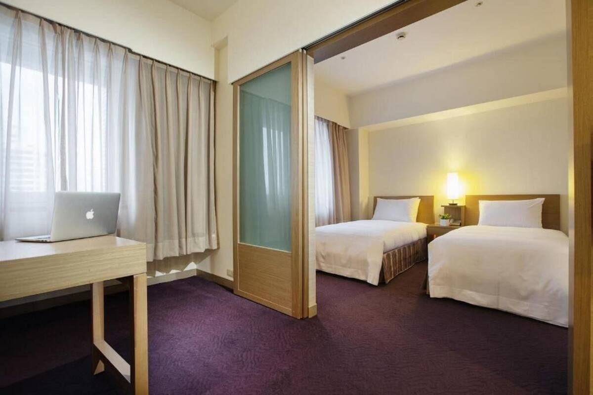 A hotel room with two beds, a desk with a laptop, and a sliding door.
