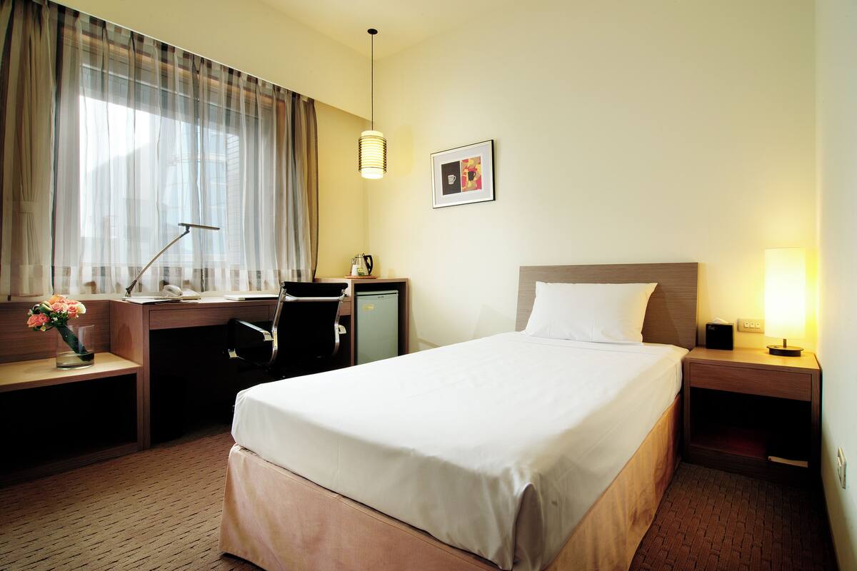 A hotel room with a bed, desk, chair, and a small table with a vase of flowers.