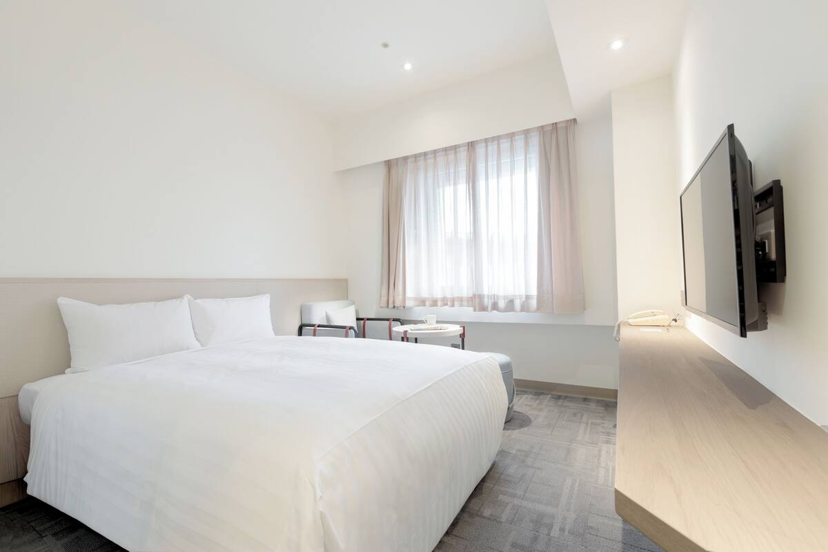 A modern hotel room with a large bed, a small desk, a chair, a TV, and a window with curtains.