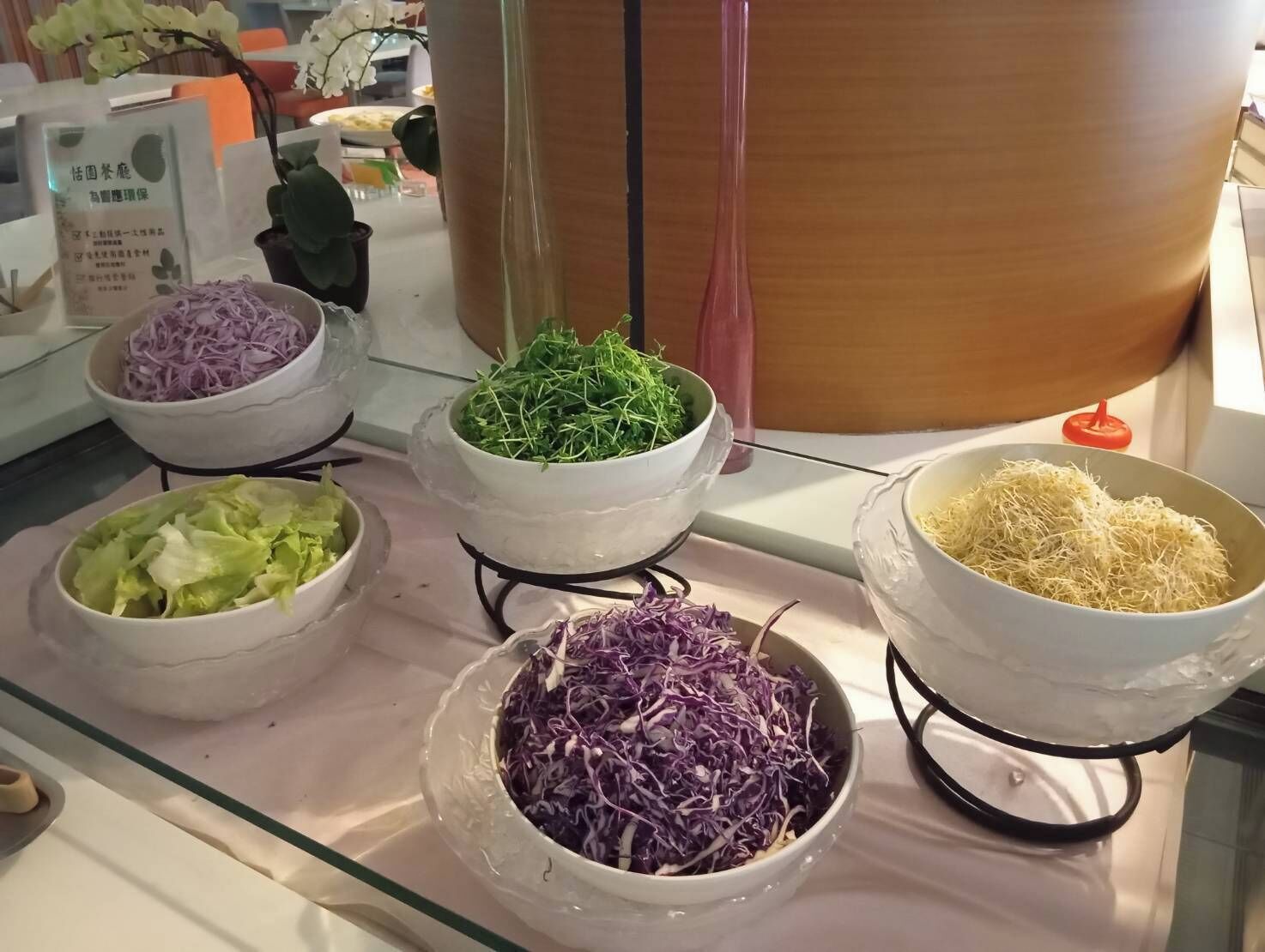 A buffet table with various food items including shredded purple cabbage, green leafy vegetables, shredded white cabbage, and shredded white noodles.