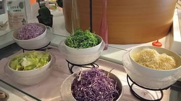 A buffet table with various food items including shredded purple cabbage, green leafy vegetables, shredded white cabbage, and shredded white noodles.
