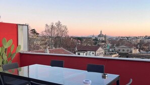 A rooftop terrace with a glass table and chairs, a cactus plant, and a view of a cityscape with historical buildings. - Farnesina Rooftop Terrace (Roma)
