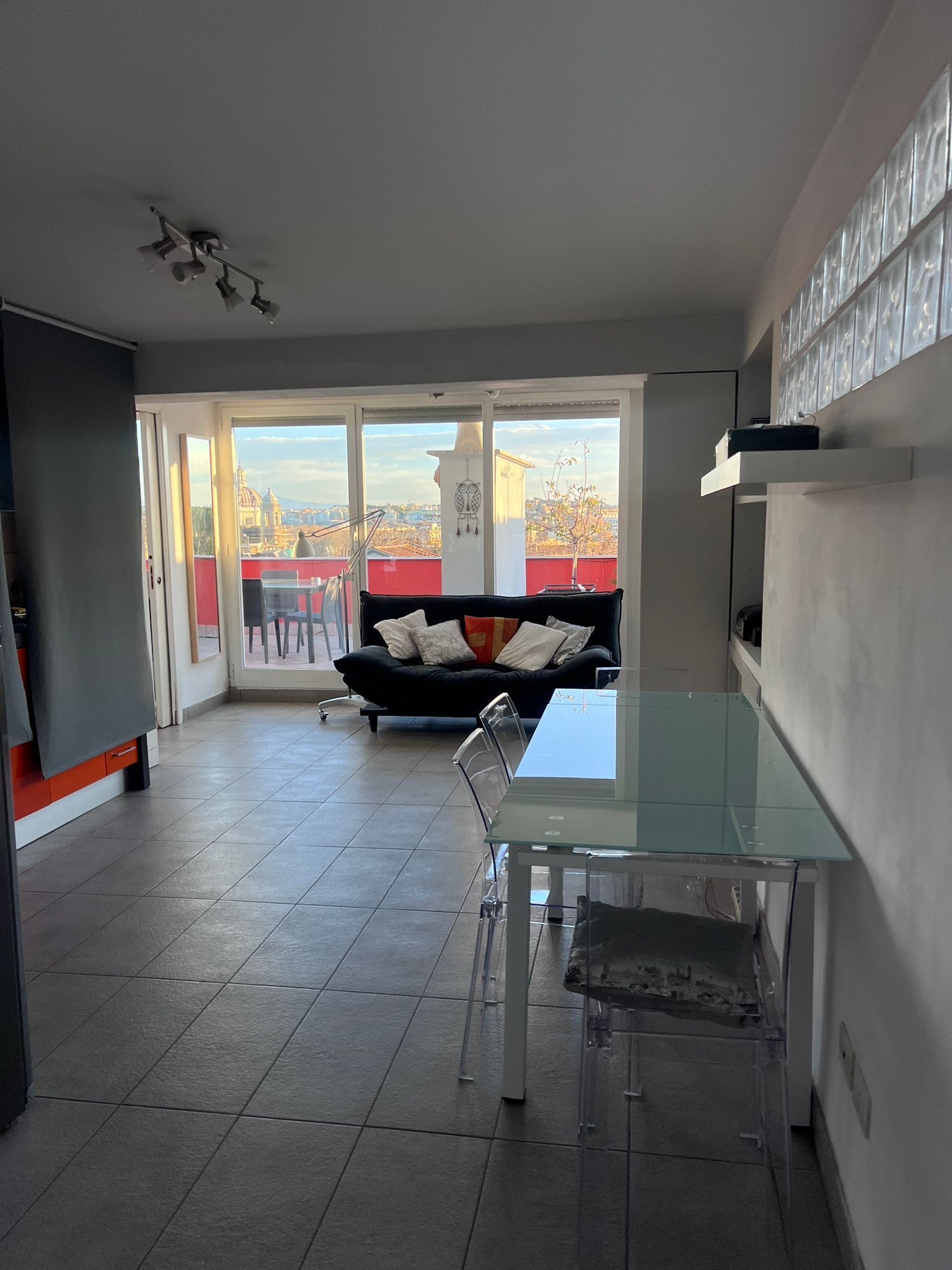Een moderne keuken met een glazen eettafel en stoelen, een zwarte bank met kussens, en grote ramen met uitzicht op de stad.
