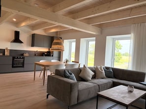 Un soggiorno moderno con un divano, un tavolino da caffè e un'area pranzo con tavolo e sedie. È presente una zona cucina con pensili e un fornello. La stanza ha ampie finestre e un soffitto in legno.