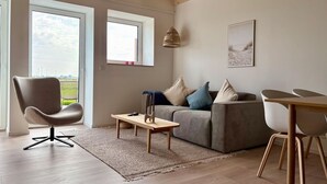 A modern living room with a grey sofa, a wooden coffee table, and a grey armchair. There are two windows with views of wind turbines and a grassy field.