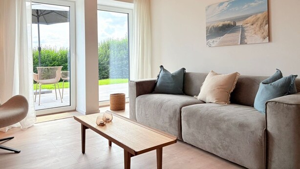 A living room with a grey sofa, a wooden coffee table, and a view of a garden through large windows.
