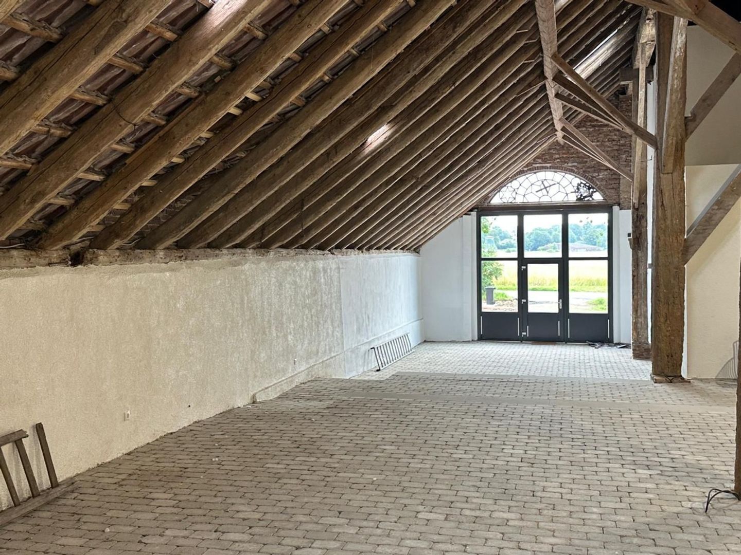 Ein Dachzimmer mit freiliegenden Holzbalken, Steinboden und einem großen Fenster mit Blick auf Grünfläche.