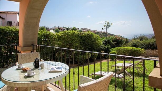 Una mesa de vidrio con sillas en un balcón que da a un jardín y edificios lejanos.