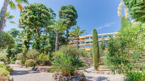Un jardin avec différents arbres et arbustes, un chemin de gravier et un bâtiment en arrière-plan.