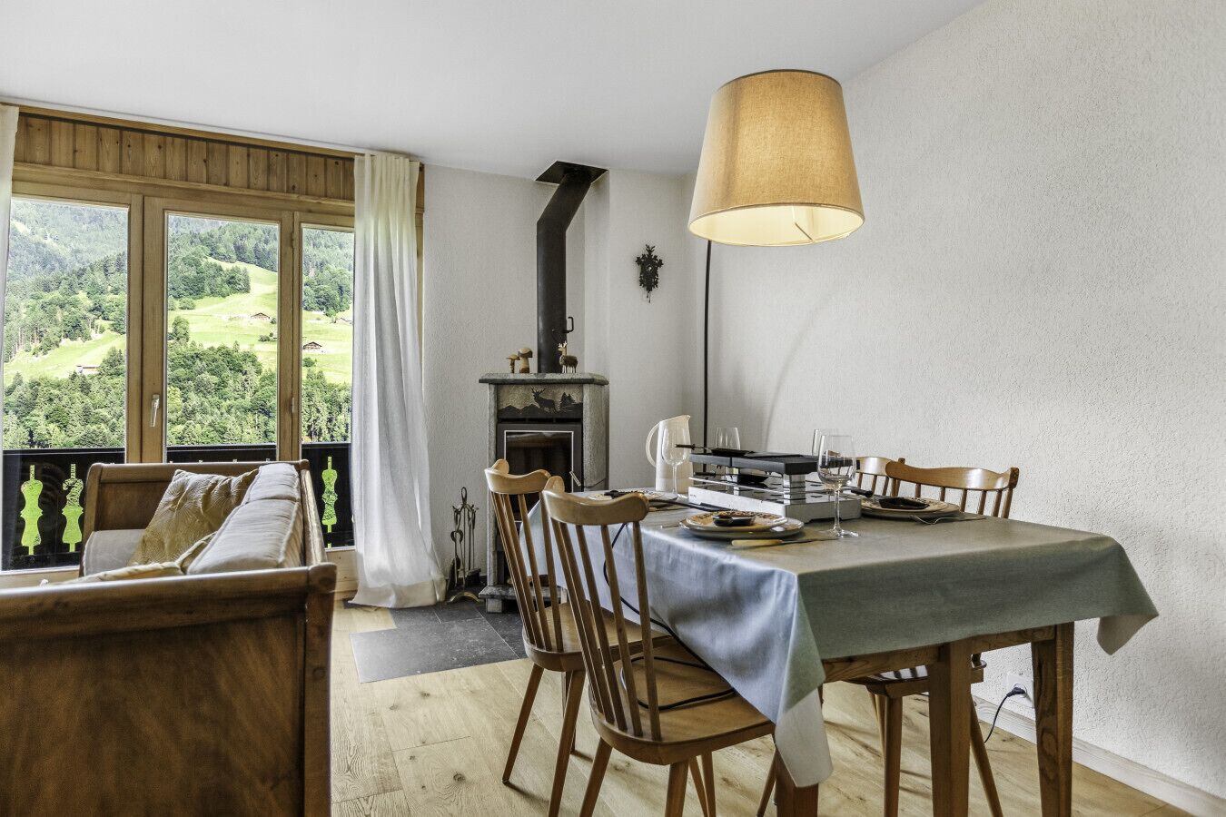 A dining area with a table set for two, wooden chairs, a fireplace, and a view of a green landscape through large windows.