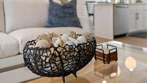 A modern living room with a glass coffee table, a decorative black basket with items, a white sofa, and a kitchen area in the background.