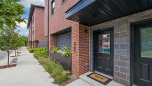 A brick building with a black door, a doormat, and a sidewalk with trees and plants.