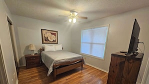 A bedroom with a wooden bed, a nightstand, a lamp, a ceiling fan, a window with blinds, and a TV on a wooden stand.