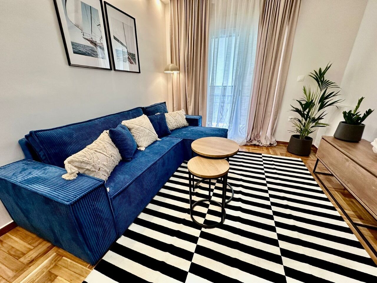 A modern living room with a blue sofa, two round wooden tables, a black and white striped rug, and a wooden cabinet.