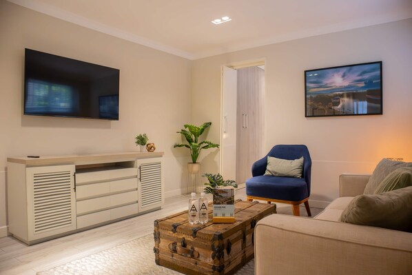 A living room with a flat-screen TV, a blue armchair, a wooden coffee table with bottles, and a painting on the wall.