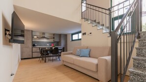 Una sala moderna con un sofá, un comedor y una cocina al fondo. Se observa una escalera con barandilla metálica que sube a un nivel superior.