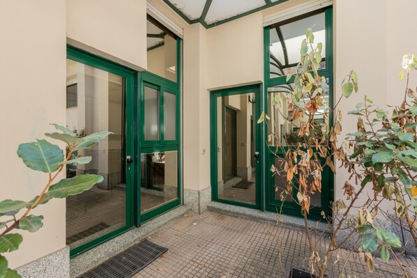 A building entrance with green-framed glass doors, a tiled floor, and a small plant in the foreground.