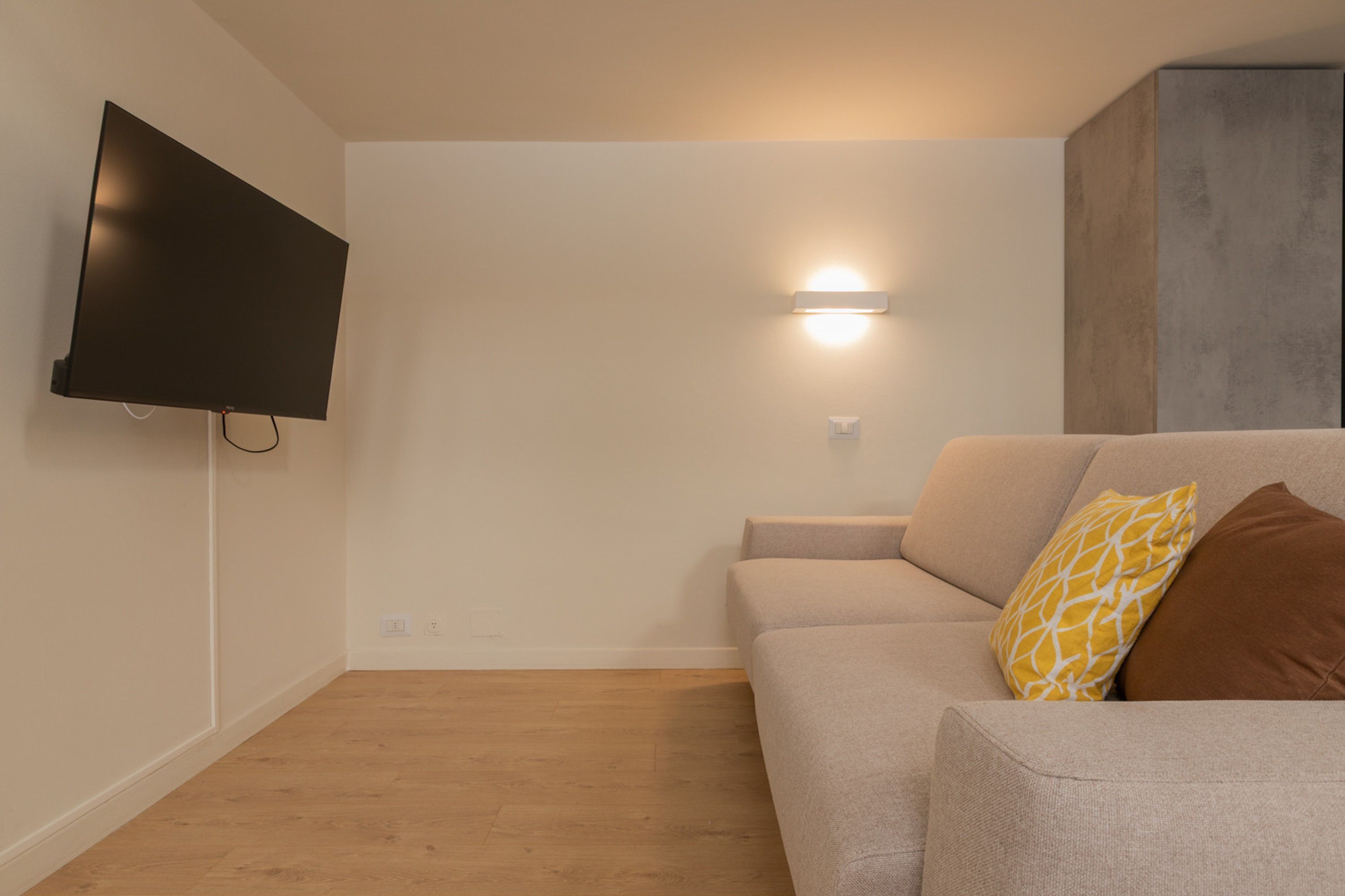 Un salon moderne avec un téléviseur à écran plat fixé au mur, un canapé avec des coussins et un luminaire fixé au mur.