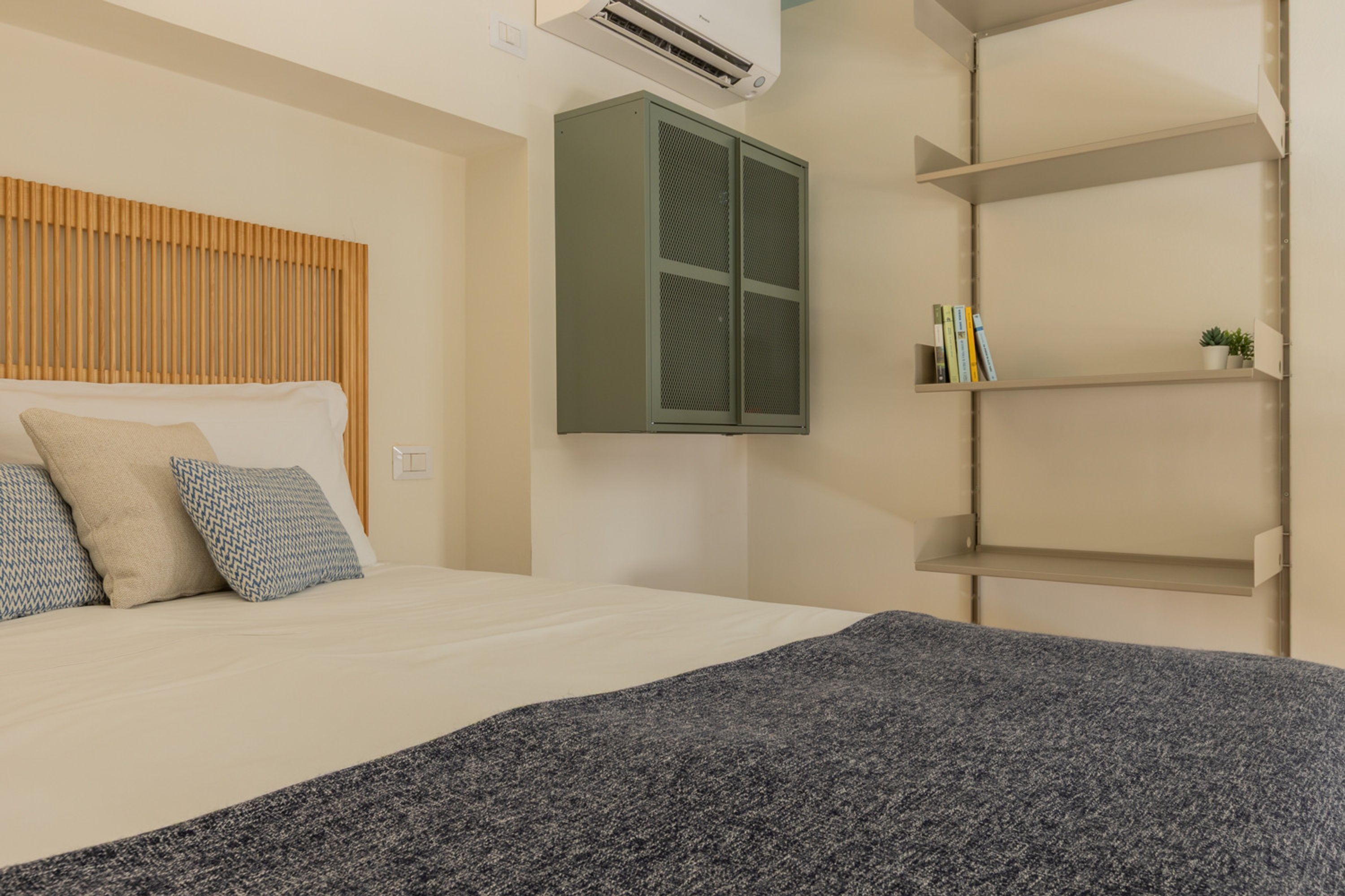 Une chambre à coucher avec un lit, un climatiseur fixé au mur, une armoire verte et une étagère avec des livres et une plante.