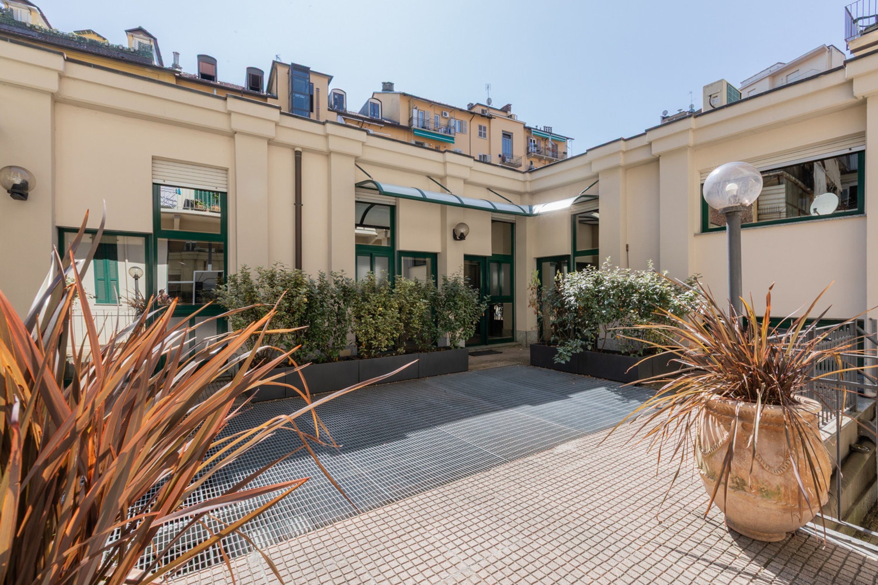 A courtyard with tiled flooring, potted plants, and a building with green-framed windows and doors.