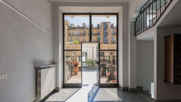 A hallway with a glass door leading to an outdoor area with potted plants and a view of buildings. - Orange Loft by Wonderful Italy (Turin)