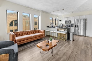 A modern kitchen with stainless steel appliances, a dining area with white chairs, and a leather sofa in a bright living room with large windows.