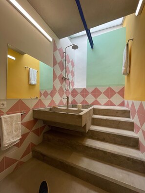 Un baño con un espejo grande, un lavamanos y una ducha. Hay escalones que llevan al área de la ducha.