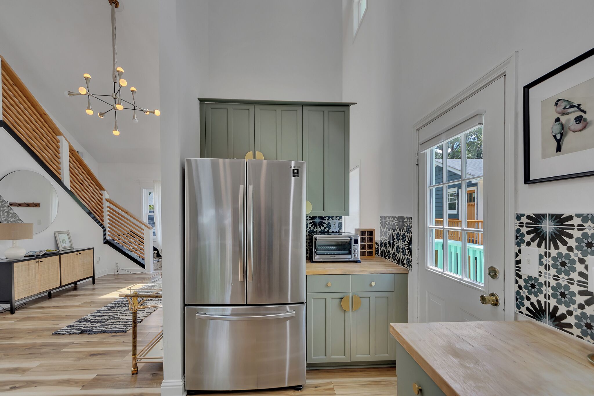A modern kitchen with a stainless steel refrigerator, a wooden staircase, and a view of the outside through a window.