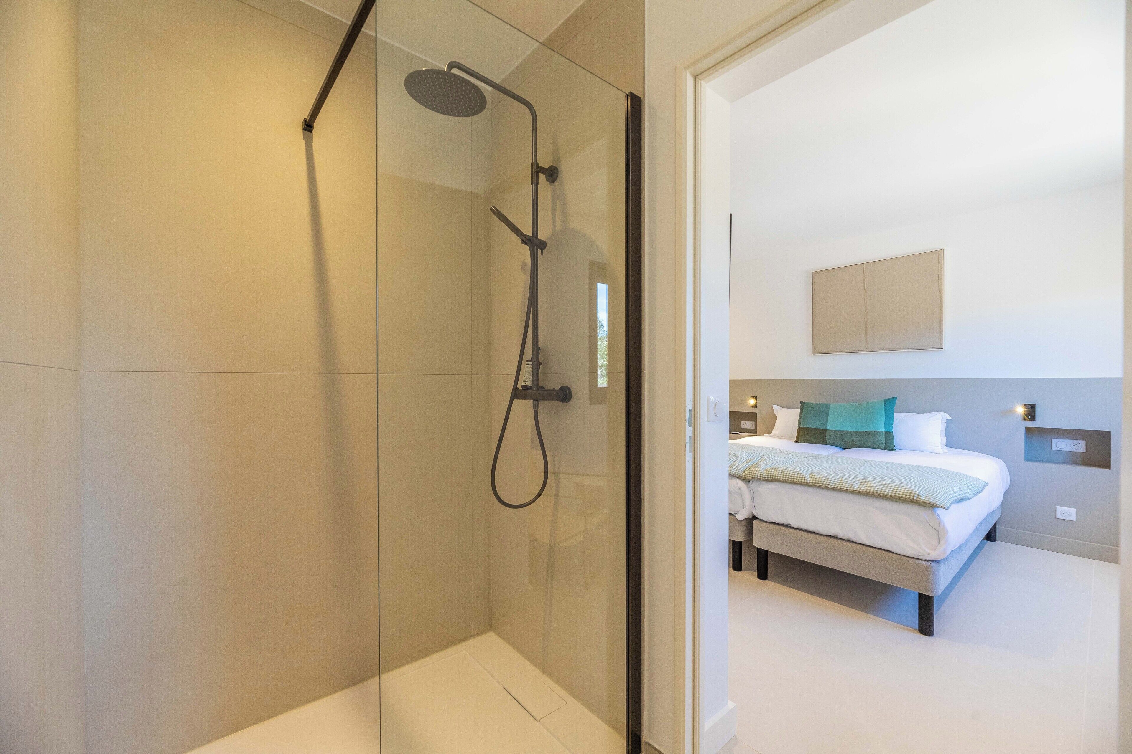 A modern bathroom with a glass-enclosed shower, a visible toilet, and a bedroom with a bed, bedside table, and a wall-mounted light fixture.