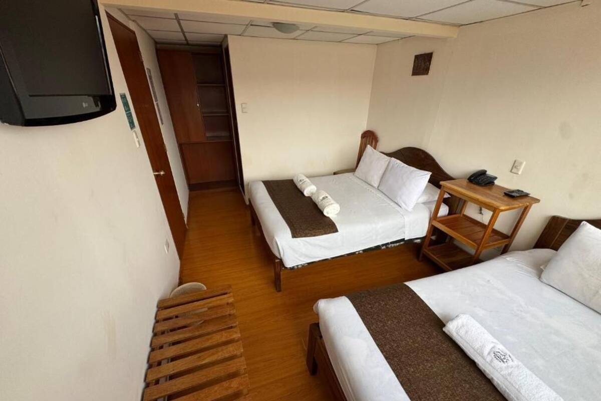 A hotel room with two beds, a wooden bench, a desk with a telephone, and a TV mounted on the wall.