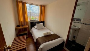 A hotel room with a bed, a bedside table, a window with curtains, and a bathroom with a sink and mirror. - Hostal El Auqui (Cusco)