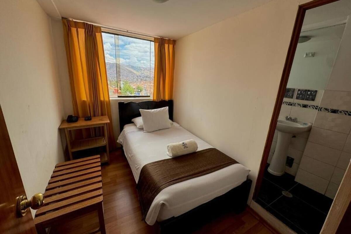 A hotel room with a bed, a bedside table, a window with curtains, and a bathroom with a sink and mirror.