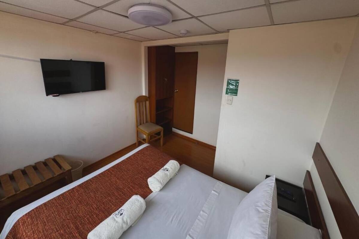 A hotel room with a bed, a TV mounted on the wall, a wooden bench, and a door leading to another room.