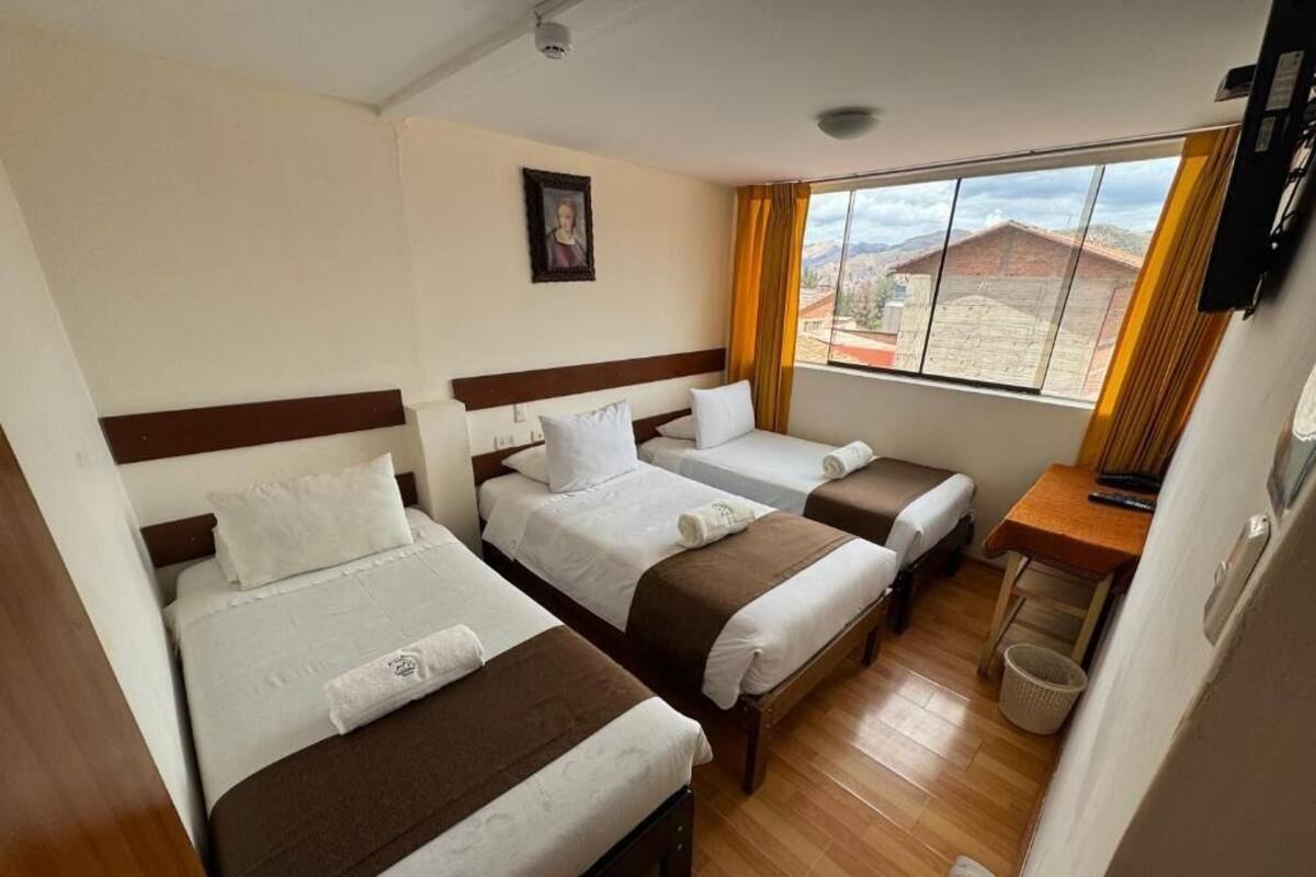 A hotel room with three single beds, a wooden floor, a small table with a chair, a TV, and a window with a view of buildings.